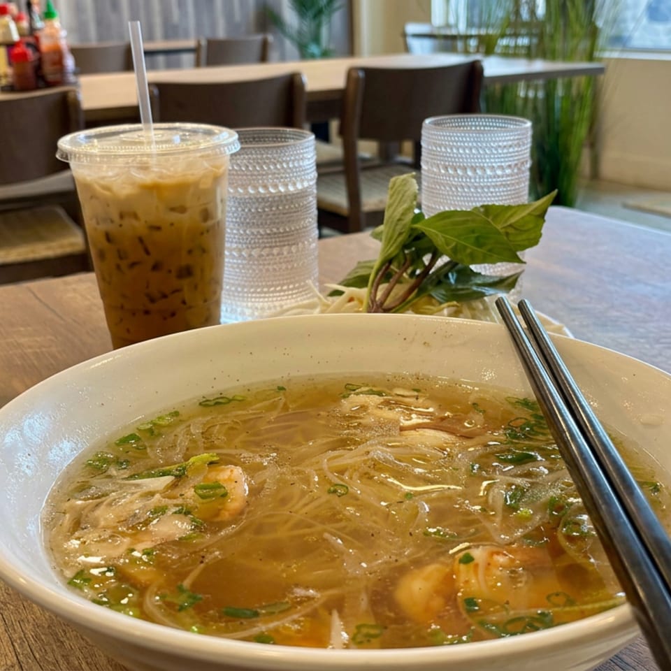 Bowl of Vietnamese noodle soup with shrimp, herbs, chopsticks, iced coffee, and fresh greens on a restaurant table.