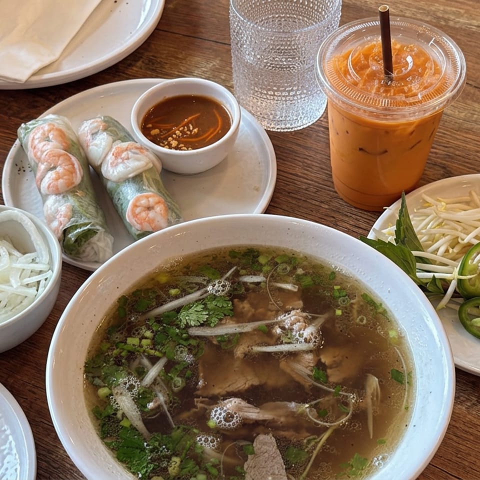 Vietnamese meal with beef pho, fresh spring rolls, herbs, bean sprouts, lime, jalapeño, dipping sauce, and iced tea on a wooden table.