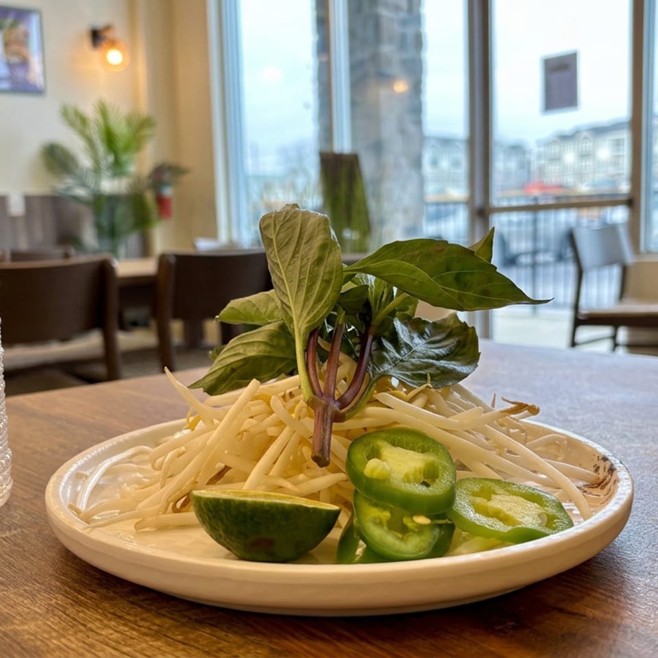 Plate of bean sprouts, Thai basil, lime wedges, and sliced jalapeños on a restaurant table between textured glasses.